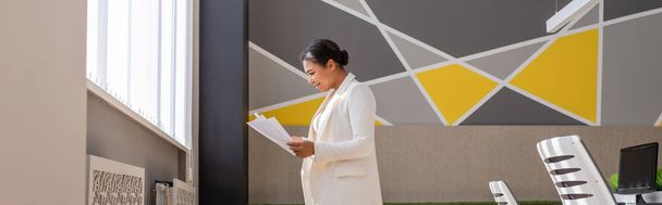 πλαϊνή άποψη της χαμογελαστή πολυφυλετική επιχειρηματίας σε λευκό σακάκι κοιτάζοντας τα χαρτιά στο σύγχρονο γραφείο, πανό - Φωτογραφία, εικόνα