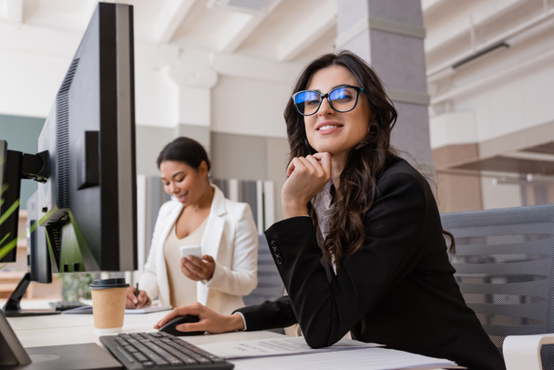 бізнес-леді в окулярах посміхається на камеру біля комп'ютерного монітора і багаторасового колеги на розмитому фоні
 - Фото, зображення