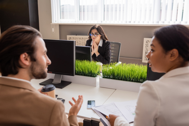 businesswoman in eyeglasses thinking near computer and interracial colleagues talking on blurred foreground - Photo, image