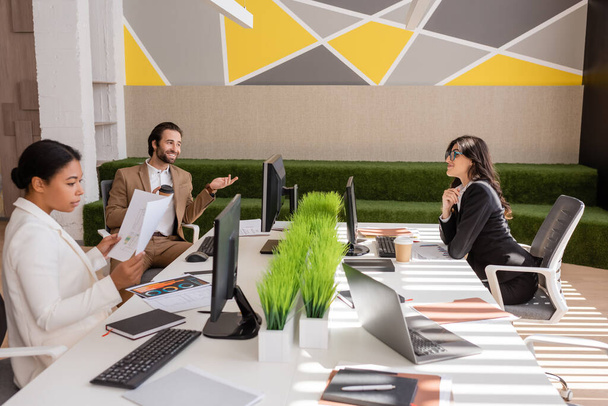 現代のオフィスでコンピュータや文書の近くで多民族の同僚と話しているビジネスマンを笑顔 - 写真・画像
