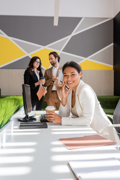 счастливая многорасовая деловая женщина с бумажной чашкой, смотрящая в камеру рядом с компьютером и коллегами с ноутбуком на размытом фоне - Фото, изображение