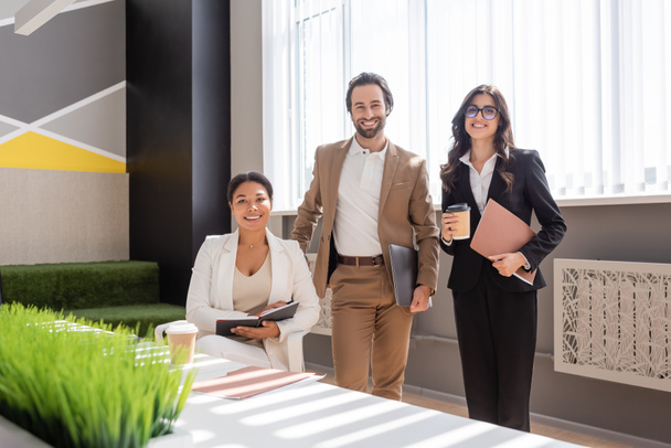 happy multiethnic business people smiling at camera near desk in modern office - Fotó, kép