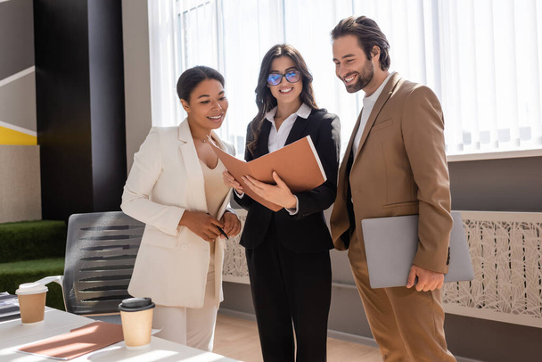 happy businesswoman in eyeglasses holding folder with documents near smiling multiethnic colleagues in office - Foto, Imagen