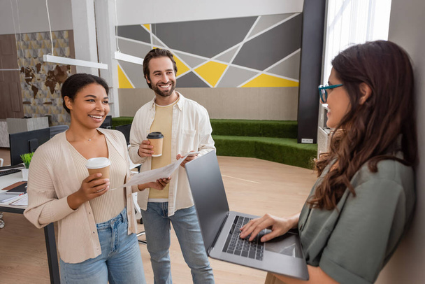 щасливі міжрасові менеджери з кавою, щоб піти дивитися на колегу, що стоїть з ноутбуком в офісі
 - Фото, зображення