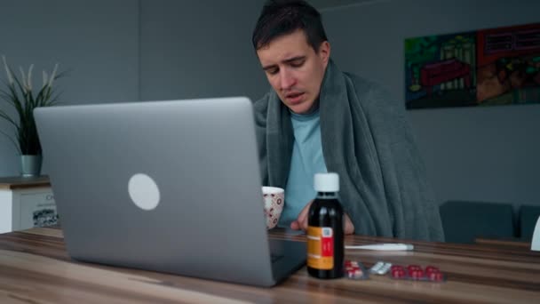 Hasta adam internette çay içip bilgisayar başında ders izliyor. Battaniyeyle kaplanmış erkek öğrenci, ilaçların yanındaki ahşap masada oturuyor. - Video, Çekim