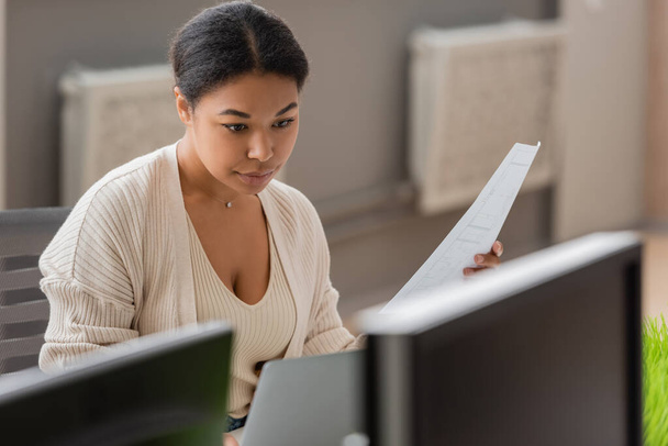 focused multiracial businesswoman holding document while working on laptop near blurred computer monitors - Photo, Image