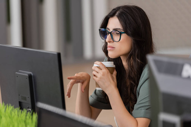 στοχαστική επιχειρηματίας με γυαλιά ηλίου κρατώντας καφέ για να πάει και δείχνοντας στην οθόνη του υπολογιστή, ενώ εργάζονται στο γραφείο - Φωτογραφία, εικόνα
