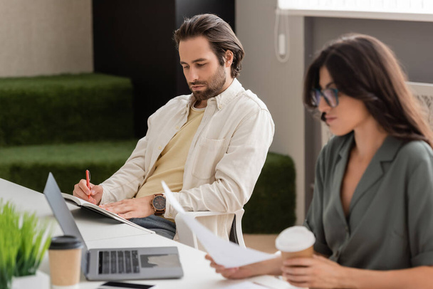 young manager with notebook and businesswoman with coffee to go sitting near laptop in office - Foto, imagen