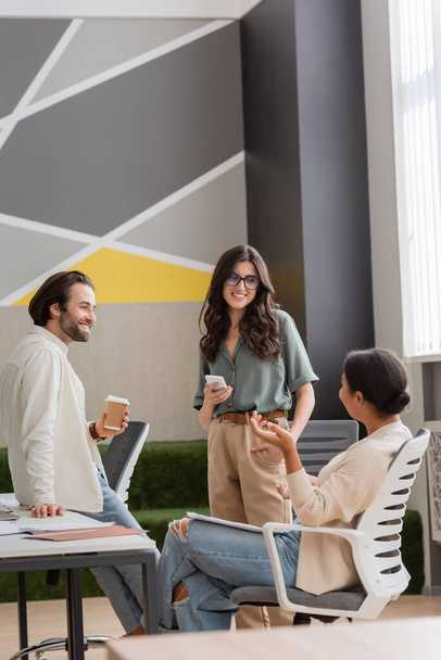 улыбающиеся менеджеры со смартфоном и кофе, чтобы пойти смотреть на многорасовых деловых женщин сидя и разговаривая в офисе - Фото, изображение