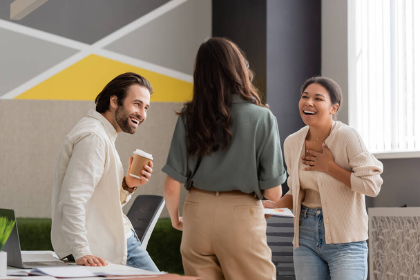ενθουσιασμένοι πολυφυλετική επιχειρηματίας αγγίζοντας το στήθος, ενώ γέλιο κοντά σε συναδέλφους στο γραφείο - Φωτογραφία, εικόνα