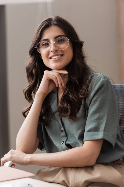 brunette businesswoman in eyeglasses holding hand near chin while smiling at camera in office - Fotoğraf, Görsel