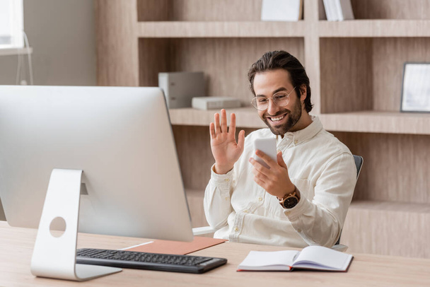 щасливий бородатий бізнесмен в окулярах махає рукою під час відеодзвінка на смартфон біля монітора комп'ютера
 - Фото, зображення
