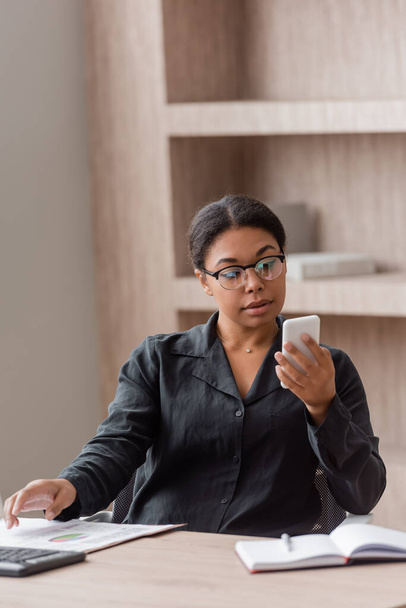 брюнетка мультирасовый менеджер в очках глядя на мобильный телефон, глядя на смартфон рядом с размытым ноутбуком - Фото, изображение
