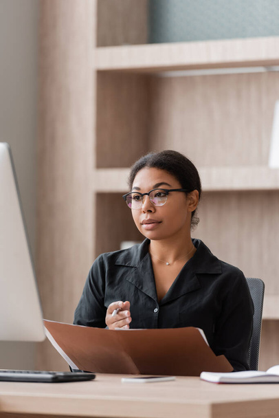 serious multiracial businesswoman in eyeglasses holding folder and looking at blurred computer monitor - Foto, immagini