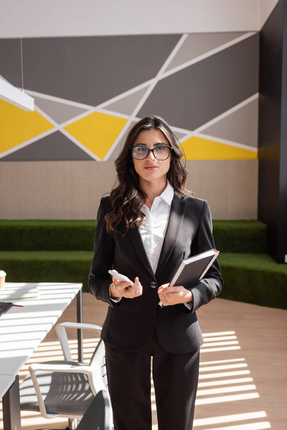 Брюнетка бизнесвумен в очках и черном блейзере стоит со смартфоном и ноутбуком в современном офисе - Фото, изображение