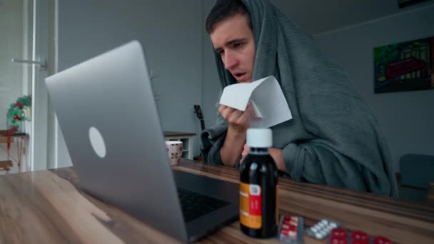 Battaniyeye sarılı bir adam ağzı peçeteyle kapatıyor. Hasta adam laptopta çalışmaya çalışıyor. Tahta masada ilaçların yanında oturuyor. Yavaş çekimde yakın çekim. - Video, Çekim