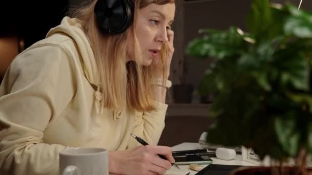 Mulher usando fones de ouvido sentado à mesa no quarto escuro e usando laptop. A trabalhar até tarde no escritório. Freelancer feminino trabalha em casa escritório à noite, use laptop - Filmagem, Vídeo