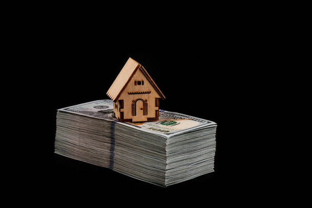 Casa de madera simbólica y una gran pila de billetes de 100 dólares americanos sobre un fondo negro - Foto, imagen