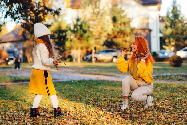 Счастливая мать и ребенок бросают осенние листья в парк. Мама и дочь веселятся вместе. Осенняя мода, стиль жизни и отдых. Семья наслаждается осенними каникулами. Стильная семья на осенней прогулке. - Фото, изображение