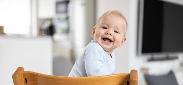 Ευτυχισμένο βρέφος κάθεται σε παραδοσιακό σκανδιναβικό σχεδιαστή ξύλινη υπερυψωμένη καρέκλα και γελώντας δυνατά στο σύγχρονο φωτεινό σπίτι. Χαριτωμένο χαμόγελο μωρού - Φωτογραφία, εικόνα