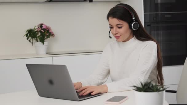 Brunette woman in headset works online on Notebook in kitchen. Young freelancer sits at table turning to look in camera smiling slow motion - Imágenes, Vídeo