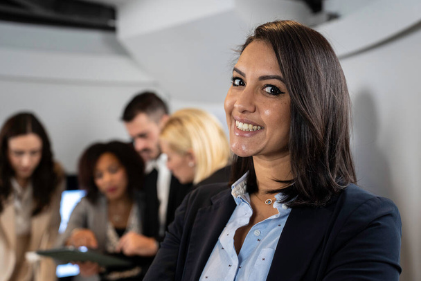 Mulher de negócios marroquina sorridente trabalhando dentro do escritório moderno com a equipe multiétnica por trás dela Conceito de liderança e trabalho em equipe atingindo metas Foco nos olhos - Foto, Imagem