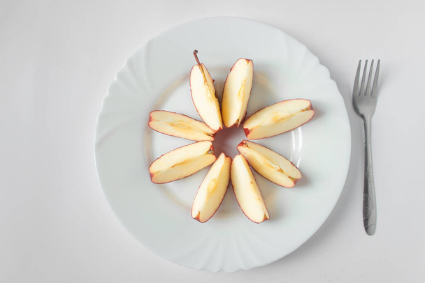 Яблоко разрезают на восемь ломтиков на белой тарелке с серебряной вилкой. Белый фон. Готов к здоровому питанию. День поста, диета и фруктовая концепция. - Фото, изображение