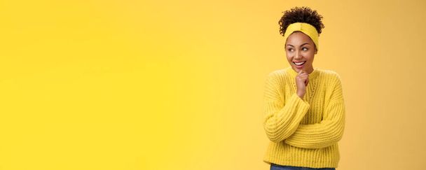感動的な驚きの幸せな魅力的なアフリカ系アメリカ人の巻き毛の女の子のヘッドバンドセーターギギング驚きの表情を左に触れる顎思慮深い笑い面白いシーン、立って黄色の背景. - 写真・画像