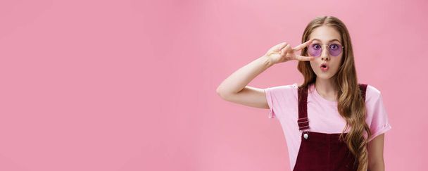Životní styl. Portrét stylové energické dívky s jizvou na ruce ukazující mírové znamení přes oko nosí chladné sluneční brýle a manšestrové kombinézy skládací rty, takže roztomilá tvář fotografování proti růžové - Fotografie, Obrázek