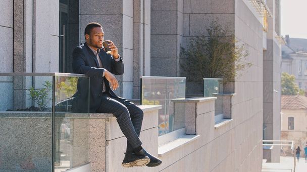 オフィス街のビルの近くの屋外に座っているアフリカ系アメリカ人の男性ビジネスワーカーは、仕事のお茶を飲むことを考えて仕事休憩をお楽しみください。外でコーヒーを飲む民族実業家 - 写真・画像
