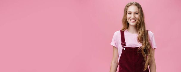 Lifestyle. Waist-up shot of kind friendly-looking pleasant young female student with long wavy fair hair in trendy overalls wearing makeup smiling joyfully and gazing at camera with good mood over - Photo, Image