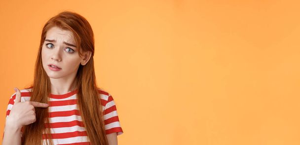 Offended innocent timid redhead girl hurt feelings, hear upsetting accusations, pointing herself frowning, look insulted, grimace frustrated, stand uneasy bothered orange background. - Photo, Image