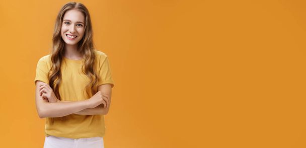 Életstílus. Kedves és barátságos fiatal lány portré, alkalmi pólóban, hullámos természetes hosszú hajjal, kéz a kézben, széles mosollyal és gondtalanul a kamera előtt narancssárga felett. - Fotó, kép