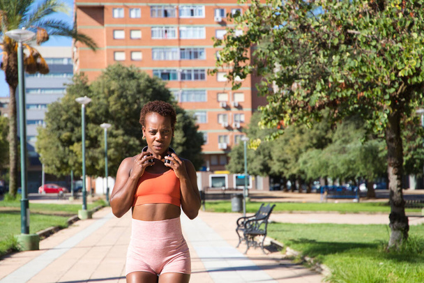 Νεαρή και όμορφη Αφρο-Αμερικανίδα με σμιλεμένο σώμα στο πάρκο, ντυμένη με πορτοκαλί μπλούζα και καλσόν. Έννοια του αθλητισμού και της υγείας. - Φωτογραφία, εικόνα