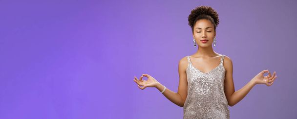 Vreedzaam zelfverzekerd opgelucht Afrikaans-Amerikaanse jonge vrouw elegante zilveren jurk kalmeren mediteren nauwe ogen ademhaling praktijk staande nirvana lotus yoga pose vinden zen, blauwe achtergrond. - Foto, afbeelding