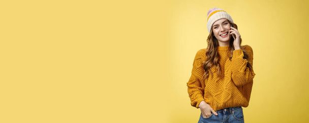 Καλαίσθητο κομψό ευρωπαϊκή γυναίκα φορώντας πουλόβερ καπέλο γέρνει το κεφάλι φλερτ χαμογελαστός μιλώντας smartphone καλώντας φίλο διαβούλευση μέσω κινητού τηλεφώνου, στέκεται ευτυχώς casual θέτουν κίτρινο φόντο. - Φωτογραφία, εικόνα