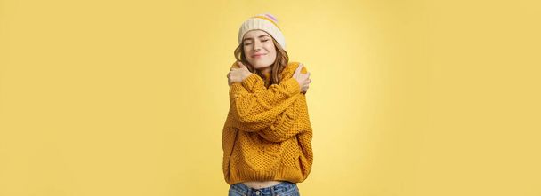 アップビート魅力的な陽気な若い女性は幸運な幸せを感じます新しい暖かい快適なセーターのように自分自身を受け入れ肩を傾けます近い目は広く巨大な自分の体を笑顔,黄色の背景. - 写真・画像