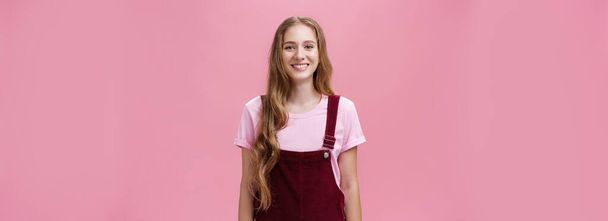Lifestyle. Portrait of charming slim young girl with long fair hair in corduroy overalls smiling joyfully standing upright with positive grin posing against pink background happy and friendly. - Photo, Image