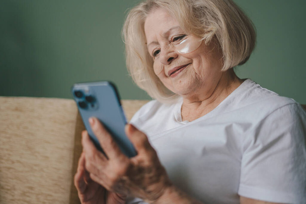古い女性は、ビデオカメラを介して電話で会話をし、ソファの上に座っている。携帯電話通信。現代のコミュニケーション。高齢者 - 写真・画像