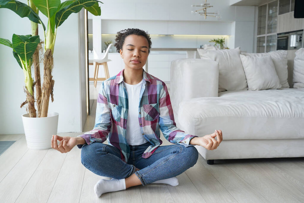 Mieszana dziewczyna rasy praktykuje jogę, medytuje, oddycha czystym powietrzem na podłodze w domu. Afroamerykanka młoda kobieta, siedząca w lotosowej pozycji z gestem mudry podczas medytacji. Zdrowy styl życia, wellness. - Zdjęcie, obraz