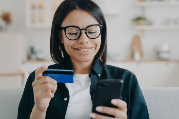 Ευτυχισμένη γυναίκα που κατέχει πιστωτική κάρτα και smartphone, χρησιμοποιεί online τραπεζικές υπηρεσίες, mobile bank app στο σπίτι. Νεαρή γυναίκα κάνει εύκολη ασφαλή πληρωμή, μεταφορά χρημάτων, ψώνια στο διαδίκτυο. Ηλεκτρονική τραπεζική. - Φωτογραφία, εικόνα
