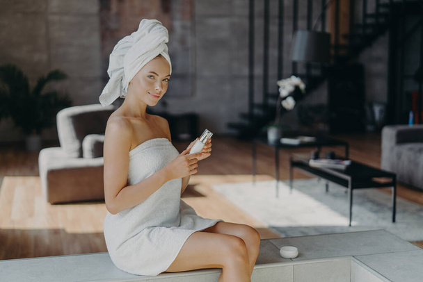 Poziomy strzał atrakcyjnej uroczej kobiety siedzi na przytulnym tle pokój stosuje balsam do ciała po kąpieli wykorzystuje produkt kosmetyczny do świecącej skóry owinięte w ręcznik przechodzi zabiegi kosmetyczne - Zdjęcie, obraz