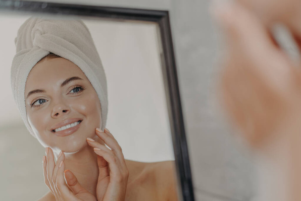 Healthy adorable young woman looks at her reflection in mirror, touches healthy glowing smooth skin, wears minimal makeup, bath towel wrapped on head after taking shower. Beauty routine concept - Photo, Image