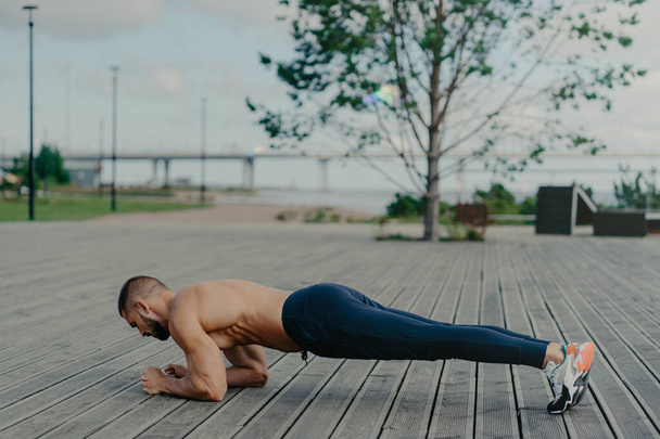Horizontal shot of sporty bearded man stands in plank pose, practices yoga outdoor and dressed in active wear, breathes fresh air. Sport, fitness and healthy lifestyle concept. Training workout - Photo, Image