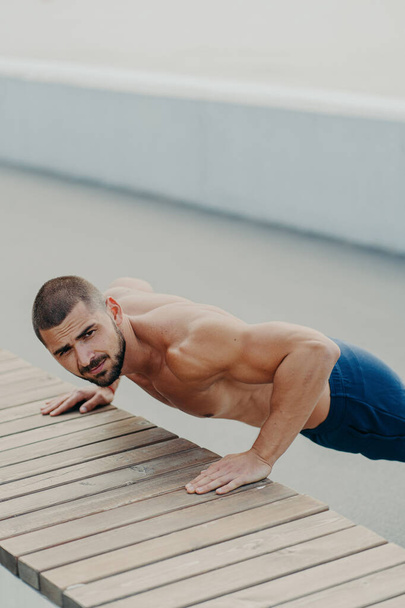 Vertical shot of muscular bearded man stands in plank pose trains chest and arms muscles has hard workout for keeping athletic body shape poses outdoor. Active lifestyle and motivation concept - Photo, Image