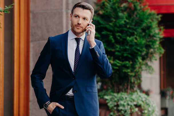 Young male CEO has telephone conversation, looks confidently into distance, dressed in elegant suit, poses outdoor, uses modern cell phone. Successful businessman has talk with business partner - Foto, Imagen