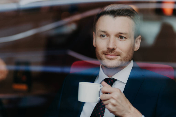 Serious concentrated unshaven male entrepreneur looks thoughfully in window of coffee shop, drinks tea, thinks about future meeting with partners, wears elegant clothing. Coffee break concept - Foto, Imagen