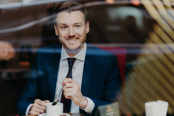 Horizontal shot of happy businessman dressed in formal clothing, drinks aromatic hot beverage, looks at window while sits in cafeteria, has pleased expression. People, rest and lifestyle concept - Foto, Imagen