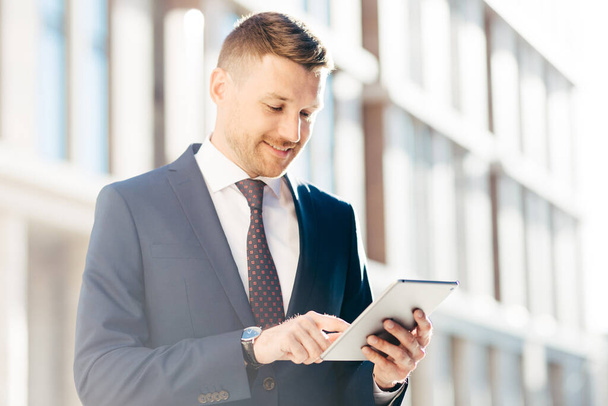 Happpy handsome male executive manager in formal elegant suit, checks income message via digital tablet, has delighted look, poses outdoor. People, business, success and professionalism concept - Foto, Imagen