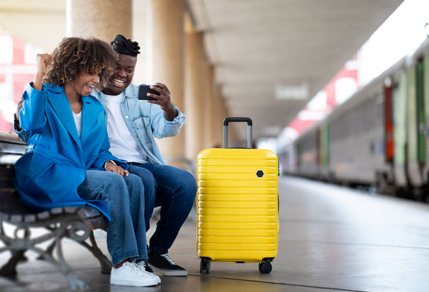 Happy Black Couple At Railway Station Celebrating Success With Smartphone, Overjoyed African American Spouses Sitting On Bench, Holding Mobile Phone And Exclaiming With Excitement, Copy Space - Photo, image